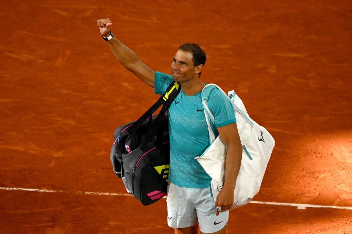 Rafael Nadal waves to the crowd while carrying his gear bags after a match on a clay court.