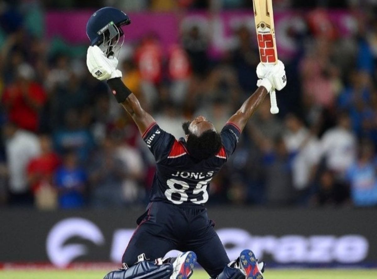 A cricket player wearing a dark jersey with the name "Jones" and the number "83" on the back, celebrates triumphantly on his knees, holding his helmet in one hand and his bat in the other, with both arms raised towards the sky.