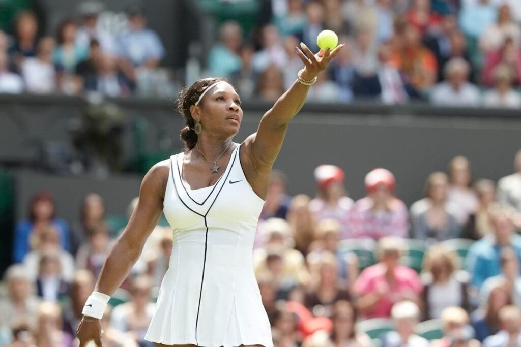 Serena Williams in a white tennis outfit, about to serve, with a crowd watching in the background.