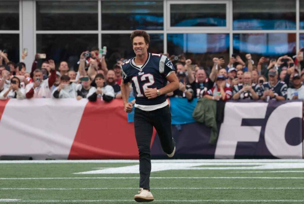 Tom Brady wearing a New England Patriots jersey, running on a football field, with fans cheering in the background.