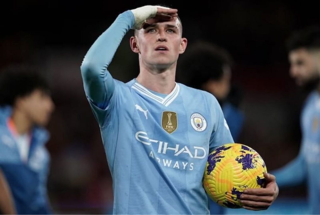 Phil Foden in a Manchester City jersey holding a soccer ball and looking into the distance with his hand on his forehead.