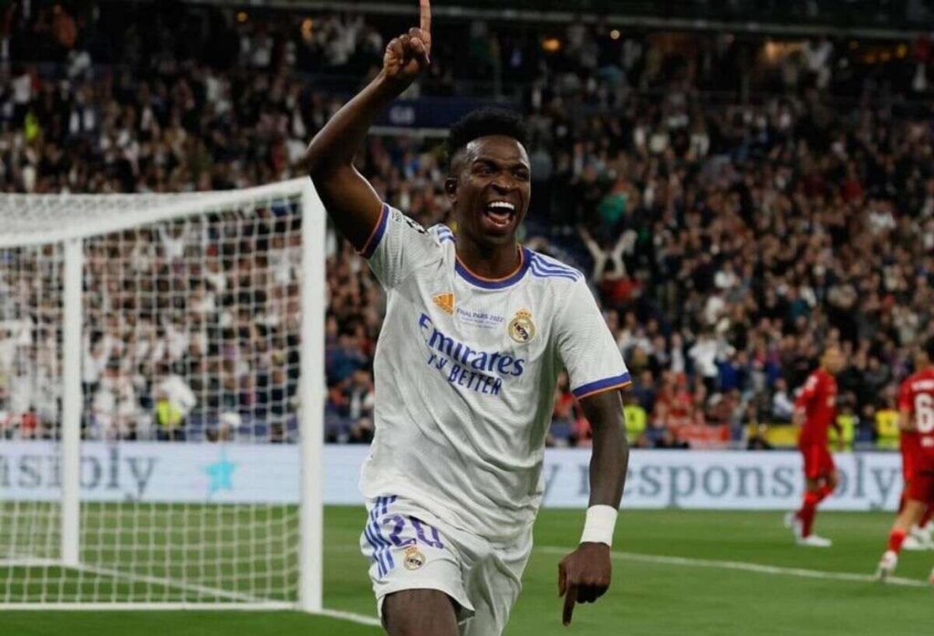 Vinícius Júnior, wearing a Real Madrid jersey, raises one finger in celebration after scoring a goal.